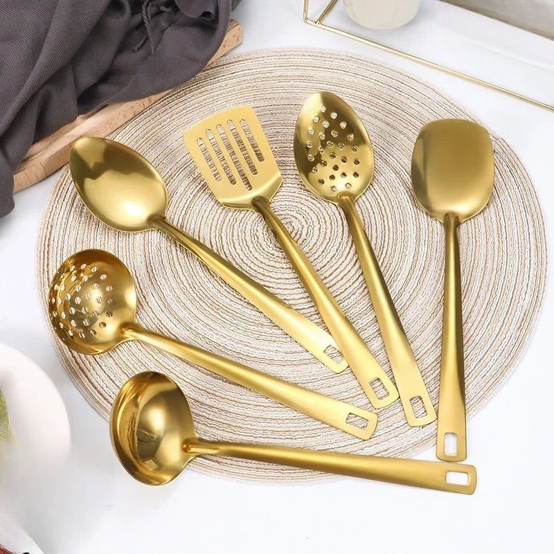 Six ustensiles de service dorés disposés sur un set de table en jonc : cuillères, louches, spatule et pelle