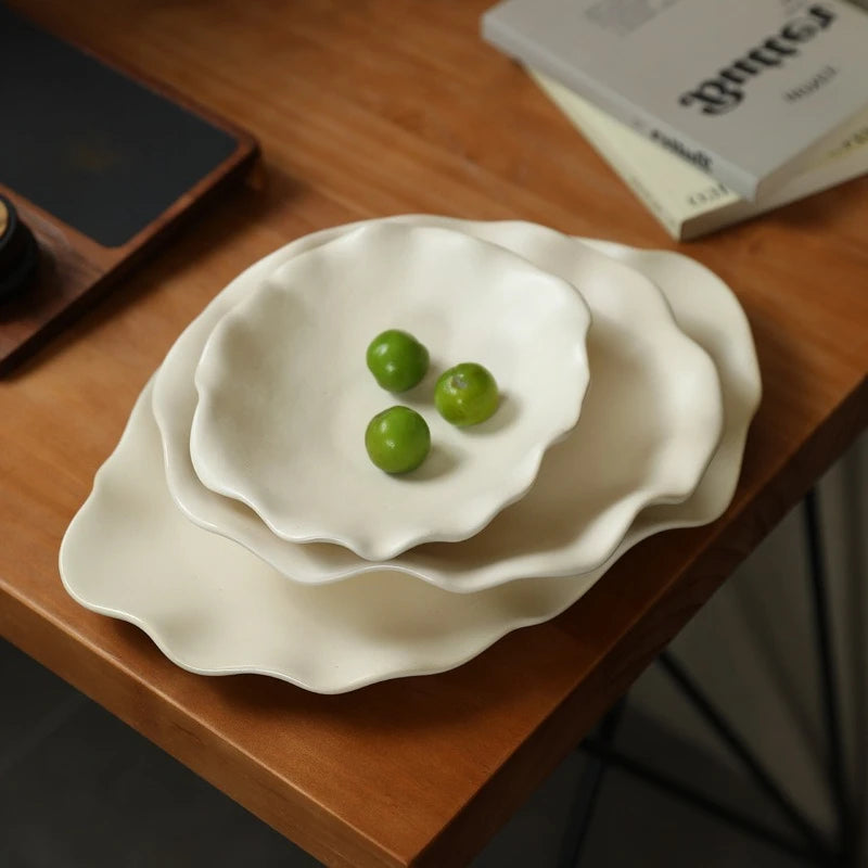 Plats en céramique crème à bord ondulé de trois tailles sur une table en bois, avec des prunes vertes
