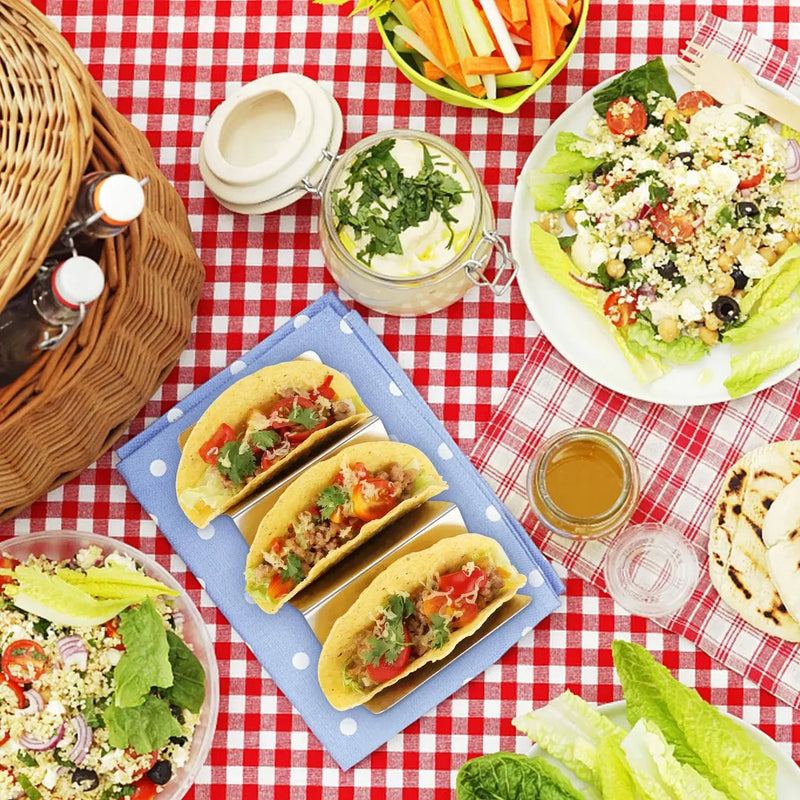Trois tacos garnis sur un support en inox posé sur une nappe à carreaux rouges avec salades et panier d'osier